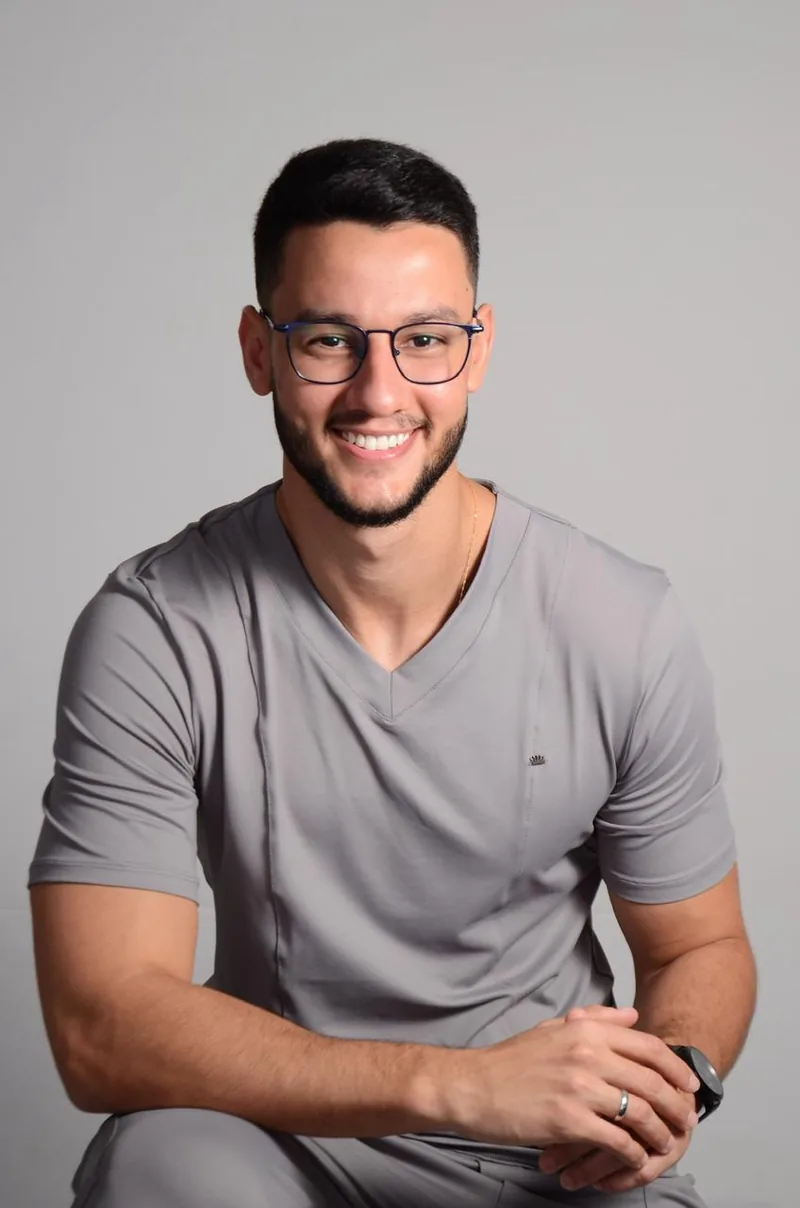 Dr. João Pedro Neves, dentista responsável pela Brasil Implantes, sorrindo, de uniforme clínico cinza e óculos